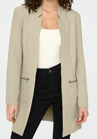Jasnobeżowa marynarka z otwartym frontem, zachowaną strukturą, dwoma przednimi kieszeniami na zamek błyskawiczny oraz teksturowaną tkaniną. Połączona z białą bluzką i czarnymi spodniami.