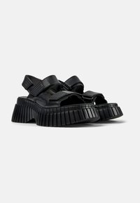 Black leather sandals with wide straps, open toe, and a distinctive ridged sole. Features Velcro closures and a chunky, textured design.