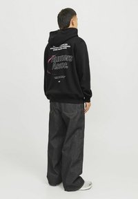 Un hoodie noir présente un grand imprimé graphique dans le dos, associé à un jean large et sombre et des baskets grises. Un design simple et décontracté.