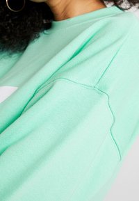 Mintgrön sweatshirt med en texturerad tyg, ribbade ärmslut och rund halsringning, samt en synlig axelsömnadsdetalj.