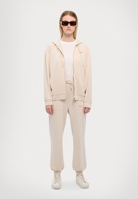 Béžový sweatsuit s kapucí na zip a kalhotami na šňůrku, s kapsami a malým logem. Doplněný bílými teniskami a tmavými slunečními brýlemi.