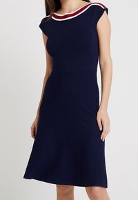Mujer que lleva un vestido azul marino hasta la rodilla con mangas japonesas y rayas blancas y rojas en el cuello.