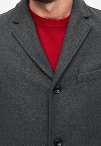 Cappotto grigio in lana con colletto a revers, due bottoni neri e una tasca sul petto, indossato sopra un maglione di lana rossa. Texture morbida e design formale.