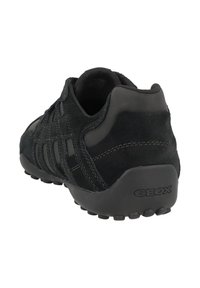 Musta mokkanahkakenkä, jossa on kumipohja, ommeltuja yksityiskohtia, pyöreä kanta ja teksturoitu pito. Brändilogo painettuna kantaan.