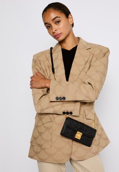 Kvinde iført en beige mønstret blazer over en sort top, beige bukser, og bærer en sort læder crossbody taske med en gylden lukning.