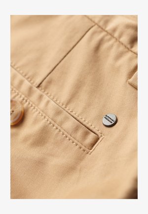 Beige stof med subtil tekstur, med en frontlomme, en plastikknap og et metalmærke med teksten "SUPERDRY PREMIUM."