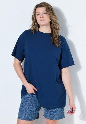 Młoda kobieta o falujących jasnobrązowych włosach nosi oversize granatowy t-shirt i teksturowane niebieskie krótkie spodenki, stojąc przy białej ścianie.