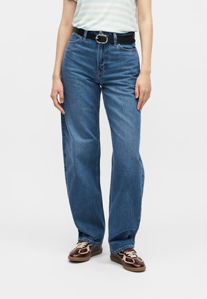 Άτομο που φοράει μπλε τζιν με ψηλή μέση και ίσια γραμμή, μαύρη ζώνη και καφέ και μπεζ αθλητικά παπούτσια, στέκεται μπροστά σε απλό φόντο.