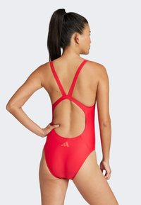 Red one-piece swimsuit with adjustable thin straps, open back design, and logo detail near the lower back. Smooth, stretchy fabric.