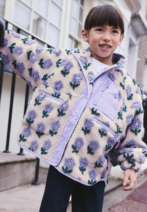 Lächelndes Kind trägt eine cremefarbene Fleecejacke mit lila Blumenmustern, steht draußen in der Nähe eines Geländers und von Gebäuden.