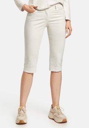 Femme portant un pantalon capri beige clair et des baskets beiges épaisses, debout avec une main dans la poche.