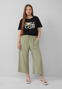 T-shirt en coton noir avec un graphique de soleil crème et du texte, associé à un pantalon large vert olive clair. Des sandales blanches à lanières complètent la tenue.