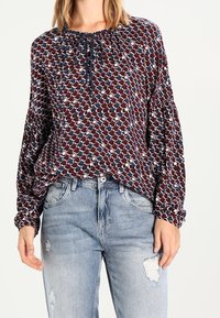 Blusa a maniche lunghe in navy e bordeaux con un motivo geometrico, caratterizzata da un colletto da annodare e maniche arricciate, abbinata a jeans chiari strappati.