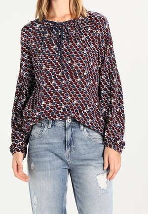 Langärmlige Bluse in Marineblau und Burgunderrot mit geometrischem Muster, mit einem gebundenen Kragen und gerafften Ärmeln, kombiniert mit hellen, lässigen Jeans.