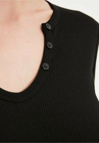 Svart ribbat ärmlöst linne med djup V-ack och fem svarta knappar längs halsringningen, som har ett mjukt, strukturerat tyg.