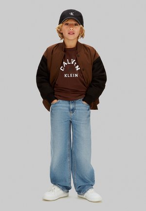 Braune und schwarze Bomberjacke, braunes "Calvin Klein" T-Shirt, hellblaue Jeans mit weitem Bein, weiße Sneakers und eine schwarze Kappe mit Logo.