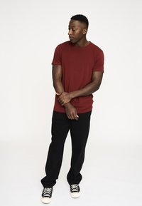 Rød crew neck t-shirt lavet af blødt stof, parret med sorte bukser og sort-hvide sneakers. Modellen står i en afslappet position.