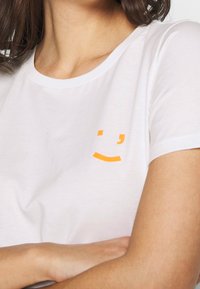 White cotton t-shirt featuring a small orange smiley face graphic on the left chest area. Round neckline with short sleeves.