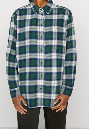 Personne portant une chemise à carreaux verte, bleue et blanche à manches longues avec un pantalon noir, debout devant un fond uni.