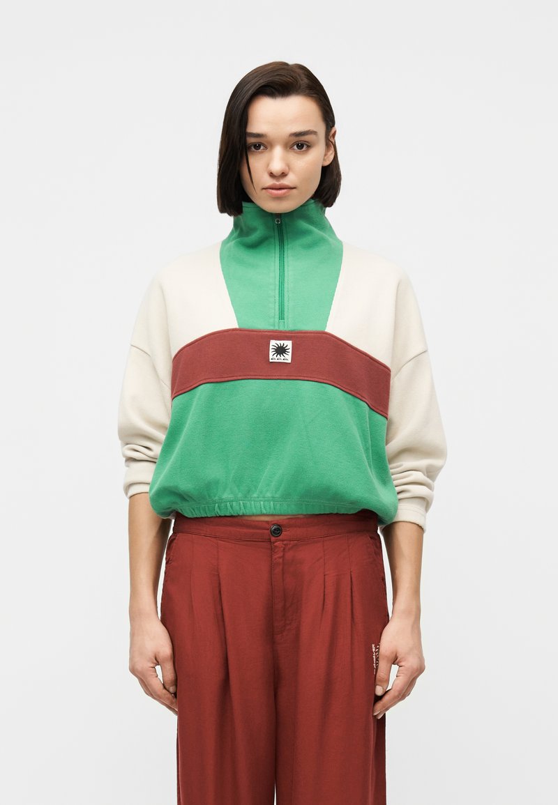 Jeune personne aux cheveux courts et noirs portant un haut zippé vert, crème et marron ainsi qu'un pantalon rouge ample, debout devant un fond uni.