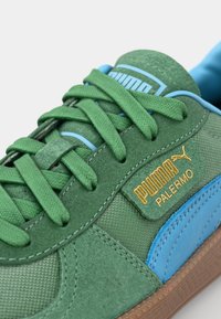 Baskets en daim vert avec des accents bleus, arborant un logo doré. Lacets plats, surface texturée et semelle en caoutchouc marron. Design sportif classique.