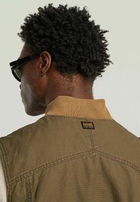 Gilet verde oliva con colletto beige a coste, dotato di un'etichetta con marchio sul retro e tessuto testurizzato. Il modello indossa occhiali da sole scuri.