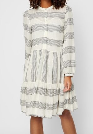 Kobieta ubrana w kremową sukienkę z czarnym, poziomym, paskowym wzorem, długimi rękawami, guzikami z przodu i marszczoną, warstwową spódnicą.