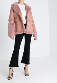 Růžová bundy z umělé kožešiny a kůže s límcem, velkými manžetami a dvěma předními kapsami. Natažena přes bílý svetřík, doplněná o černé rozšířené džíny.