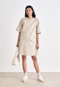 Vestido de algodão bege com mangas curtas, dois bolsos frontais e detalhe de botão nos ombros. Combinado com ténis brancos e uma pequena mala.