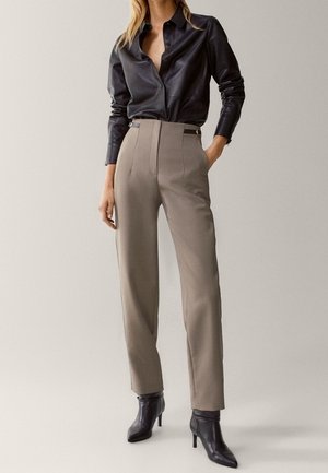 Woman wearing a black leather button-up shirt, taupe high-waist tailored pants, and black heeled ankle boots, standing with one hand in pocket.