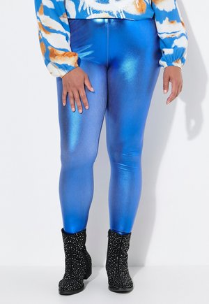 Glanzende blauwe leggings met een metallic afwerking, met een gladde textuur. Gecombineerd met zwarte enkellaarzen versierd met rhinestones.