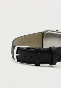 Černý kožený řemínek hodinek se stříbrnou obdélníkovou sponou, s hladkou texturou a viditelným prošitím na vnitřní straně.