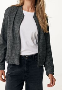 Veste bomber à motif pied-de-poule noir et blanc avec fermeture éclair à l'avant, poignets côtelés et texture lisse, portée sur un T-shirt blanc.