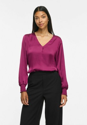 Femme aux longs cheveux foncés porte un chemisier à manches longues magenta et un pantalon noir, debout devant un fond blanc uni.