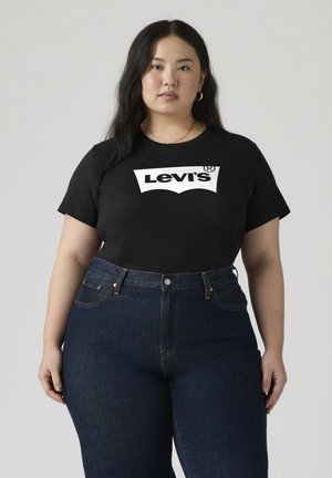 Vrouw met lang donker haar die een zwarte Levi's t-shirt en donkerblauwe, hoog getailleerde spijkerbroek draagt, staand tegen een lichte achtergrond.