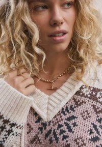 Knit sweater showcasing a v-neck design with textured cream and brown patterns. Paired with a chunky gold chain necklace.