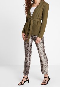 Světlý olivově zelený kabát s opaskem a texturovaným povrchem, který je spárován s béžovými a hnědými kalhotami s hadím vzorem a černými sandály na vysokém podpatku.