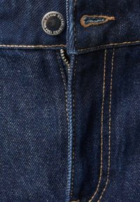 Tmavomodré džíny z denimu majú predný zips, medenú niť a kovový gombík s embosovaným logom. Detailný pohľad na pásovú časť.