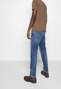 Camiseta marrón de manga corta combinada con jeans azules de pierna recta y botines marrones, sobre un fondo blanco liso.