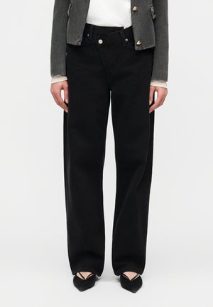 Žena v čiernych širokých džínsach s vysokým pásom, v čiernych zakončených špičkou balerínkach a krátkej tmavej bunde.