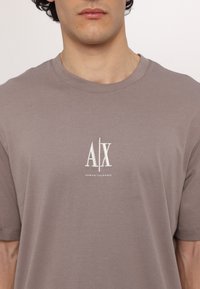 Trumpas rankovės, apvaliu kaklu marškinėliai šviesiai taupe spalvos. Panaudota balta siuvinėta logotipas "AX" ir "ARMANI EXCHANGE", centruotas ant krūtinės.