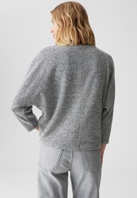 Grijze gebreide sweater met een ruime pasvorm, driekwartmouwen en een gestructureerd patroon. Draagt met lichtgrijze jeans, voorzien van achterzakken.