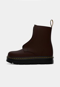Botín marrón de estilo slip-on, hecho de material suave, con una suela negra gruesa y detalles de costura amarilla. Cuenta con una lengüeta en la parte trasera.