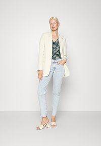 Blazer bege sobre uma blusa verde com padrão, jeans azul claro com detalhes desgastados e mules de bloco creme com ponta aberta.