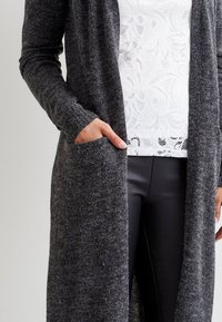 Grå, lång kofta i en strukturerad stickning med öppen framsida, ribbade ärmslut och sidofickor, lagd över en vit, mönstrad topp.