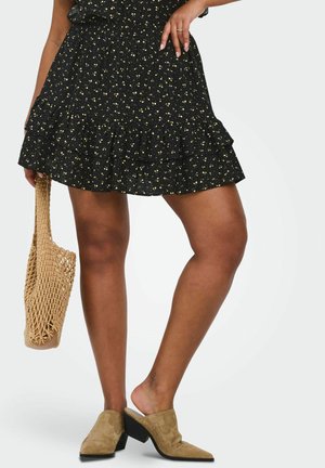 Kvinde iført sort blomsterprintet flæse nederdel, brune mule sko, holder flettet net taske, står med det ene ben let bøjet.