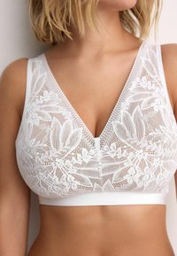 Weißes Spitzentop mit V-Ausschnitt, floralen Stickereien und einem weichen Band. Der Stoff ist leicht und hat filigrane Muster.