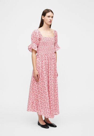 Γυναίκα που φοράει ροζ μακρύ φόρεμα με λουλούδια και φουσκωτά μανίκια, καθώς και μαύρα μπαλαρίνες, στέκεται μπροστά σε απλό λευκό φόντο.