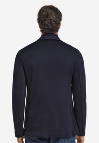 Mężczyzna w ciemnoniebieskiej marynarce z centralnym rozcięciem z tyłu, pokazany od tyłu na jednolitym białym tle.