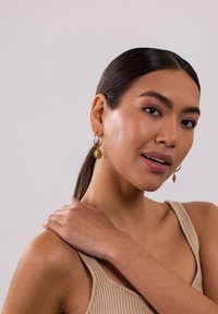 Goldene Creolen mit einem Sonnenstrahl-Anhänger und einer Muschel-Akzent, präsentiert von einem Modell, das ein beiges, geripptes Oberteil trägt, vor einem neutralen Hintergrund.